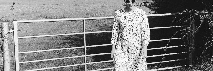 Walking along in maxi, white dress with black polka dots. Gate and field behind and sunglasses. Dress worn with socks and sneakers.